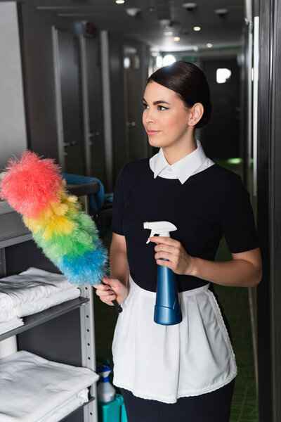 young maid in uniform holding dust brush and spray bottle 