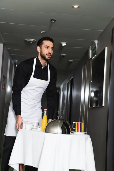 cheerful waiter in medical mask and apron delivering tray with cloche and orange juice