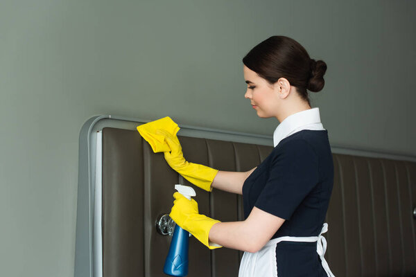 side view of young maid in rubber gloves holding spray bottle and rag while cleaning hotel room 