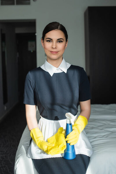smiling chambermaid in rubber gloves holding spray bottle and sitting on bed in hotel room