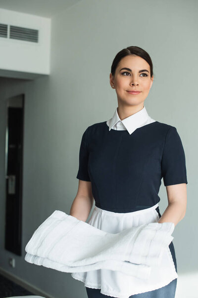 cheerful chambermaid in uniform holding clean towels in hotel room 