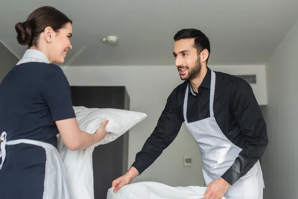 positive housekeepers changing bedding in modern hotel room