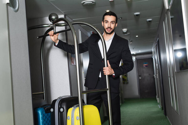bearded bellboy near metallic bell cart with luggage in hall of hotel