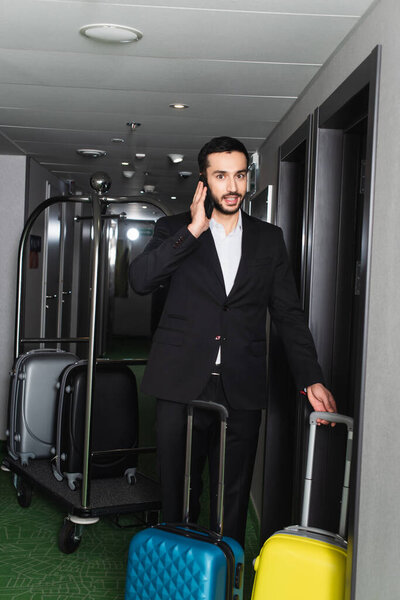 cheerful and bearded man standing with baggage and talking on smartphone in hotel corridor 