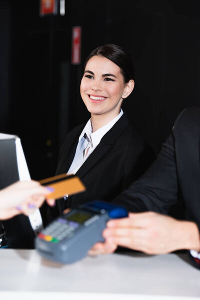 happy receptionist looking at tourist paying with credit card near colleague 