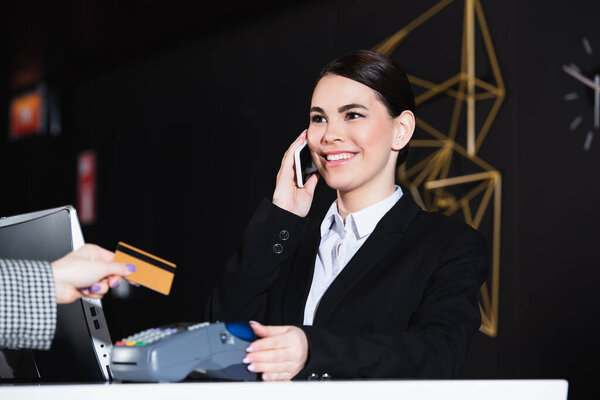 happy receptionist smiling and talking on smartphone near guest with credit card 