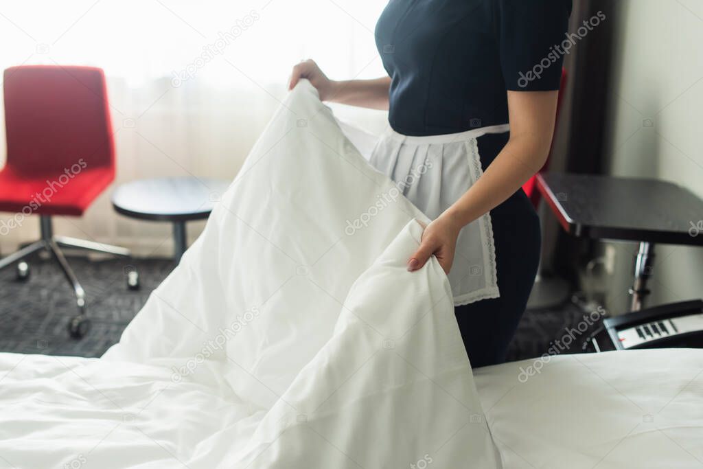 cropped view of young chambermaid in uniform changing bedding in hotel room 