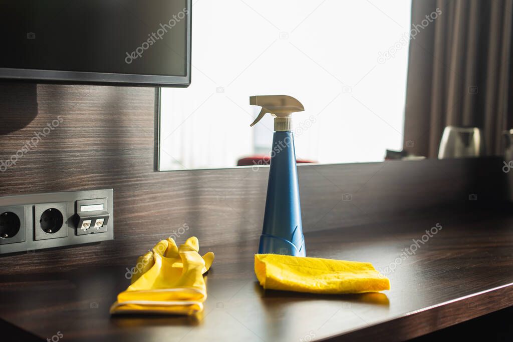 spray bottle, rag and yellow rubber gloves on wooden table in hotel room 