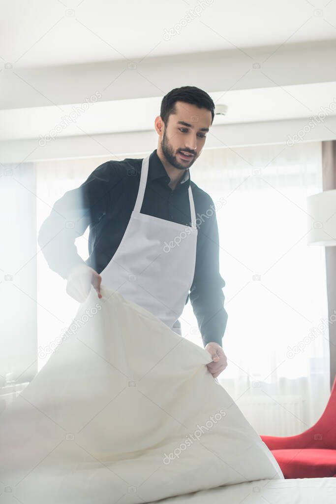 bearded housekeeper changing white bedding and smiling in hotel room