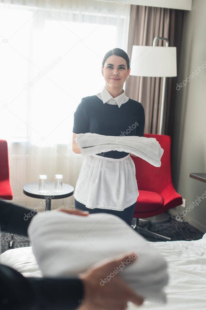 cheerful maid holding clean bed sheets near colleague with towels on blurred  foreground 