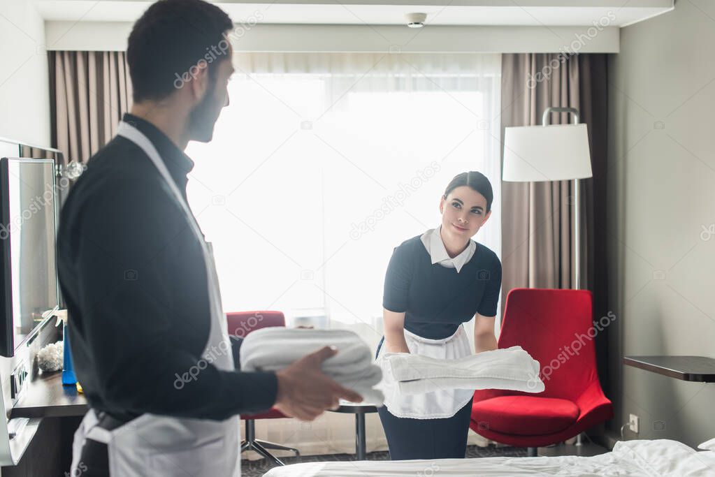 young maid holding white bed sheets near colleague with clean towels on blurred foreground 