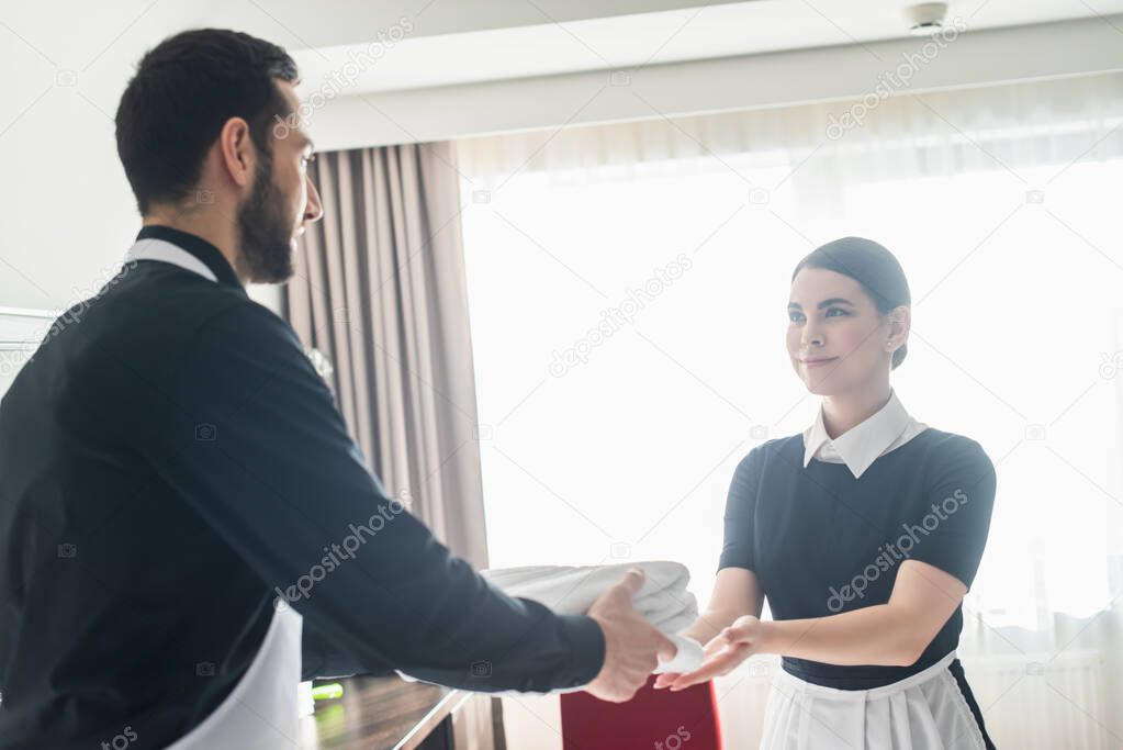 cheerful chambermaid giving towels to blurred bearded colleague 