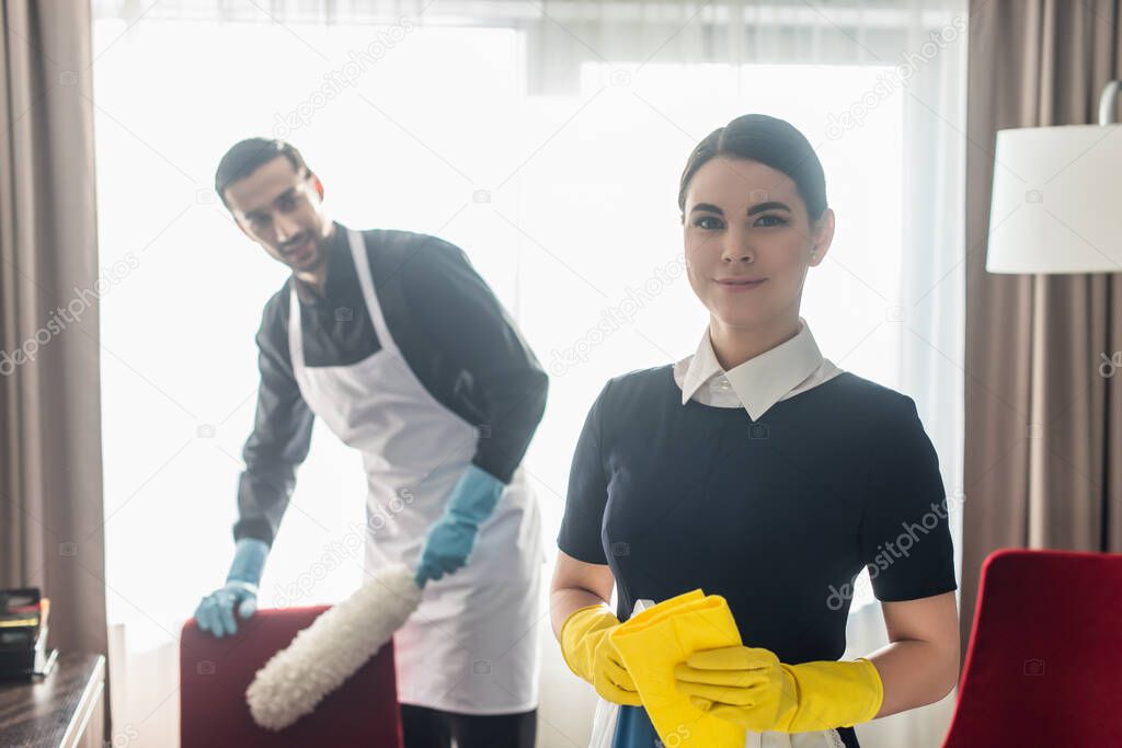 cheerful maid holding rag and smiling near blurred colleague 