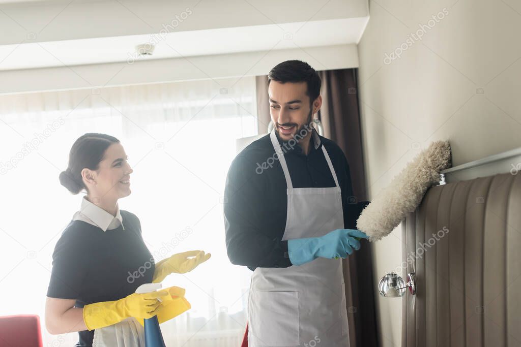 happy housekeepers looking at each other while holding cleaning supplies and cleaning hotel room