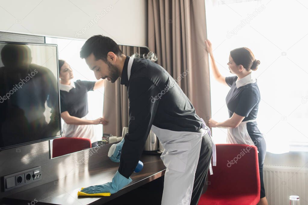 bearded housekeeper cleaning wooden surface with rag near blurred maid adjusting curtains 