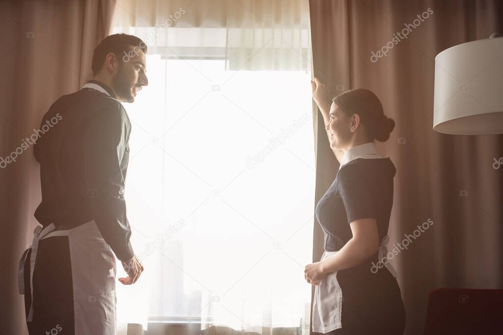 happy housekeepers adjusting curtains in hotel room 