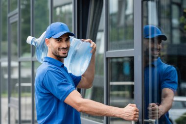 Smiling muslim courier holding bottle of water near door of building outdoors  clipart