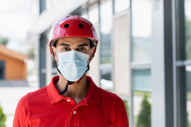 Kasklı ve tıbbi maskeli Arap kurye dışarıdaki kameraya bakıyor. 
