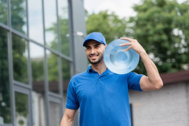 Cheerful arabian deliveryman in cap holding bottle of water outdoors  clipart