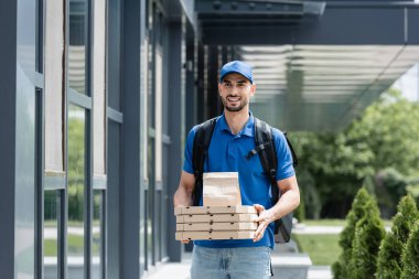 Gülümseyen Arap kurye elinde pizza kutuları ve kese kağıdıyla dışarıda. 