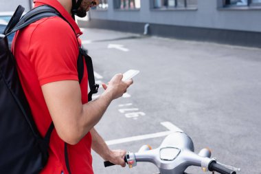 Scooter 'ın yanında akıllı telefon kullanan sırt çantalı teslimatçının görüntüsü. 