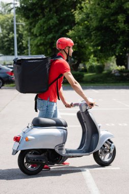 Motosikletin yanında duran termo sırt çantalı müslüman teslimatçının yan görüntüsü 