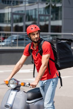 Kasklı ve termal çantalı Arap teslimatçı dışarıda scooter 'ın yanında duruyor. 