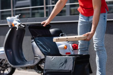 Teslimatçının termal sırt çantasının yanında pizza kutusu tutarken kısmi görüntüsü ve açık havada mobilet bulanıklığı. 