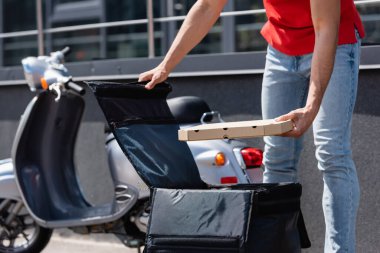 Partial view of deliveryman holding pizza box near thermo backpack and blurred scooter outdoors  clipart