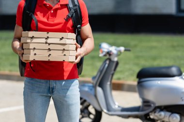 Termo sırt çantalı teslimatçının açık havada mobiletin yanında pizza kutuları tutarken çekilmiş görüntüsü. 