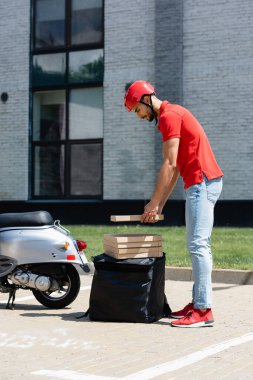Kasklı Arap kuryenin termal sırt çantası ve scooterın yanında pizza kutusu tutarken yan görüntüsü 