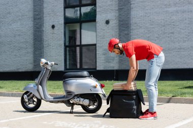 Miğferli Arap kuryenin motosikletin yanındaki termo sırt çantasından pizza kutuları alırken yan görüntüsü. 
