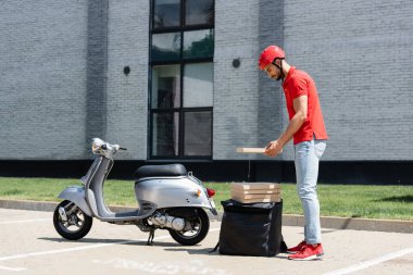 Kasklı genç müslüman teslimatçının termo sırt çantası ve scooter 'ın yanında pizza kutusu tutarken yan görüntüsü. 