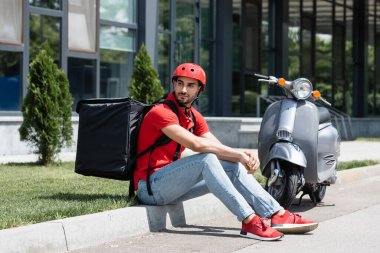 Miğferli ve termal çantalı Arap kurye dışarıda mobiletin yanında oturuyor.  