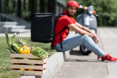 Bulanık teslimatçı ve scooter 'ın yanında taze sebzelerle dolu bir kutu. 