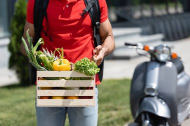 Kırmızı tişörtlü teslimatçının, bulanık scooter 'ın yanında organik sebzelerle elinde tuttuğu görüntüler. 