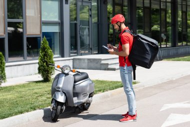 Genç müslüman teslimatçının yan görüntüsü, pano ve termo sırt çantalı dışarıda scooter 'ın yanında duruyor. 