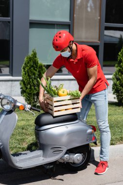 Tıbbi maskeli Arap kurye dışarıda motosikletin yanında sebzeli kutu tutuyor. 