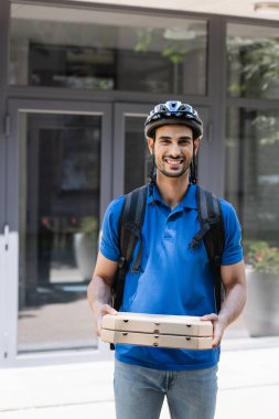 Güvenlik kaskı ve sırt çantasıyla gülümseyen Müslüman kurye pizza kutularını tutuyor ve dışarıdaki kameraya bakıyor. 