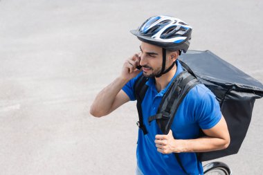 Sırt çantalı gülen teslimatçının motosikletin yanında akıllı telefondan konuşmasının yüksek açılı görüntüsü. 