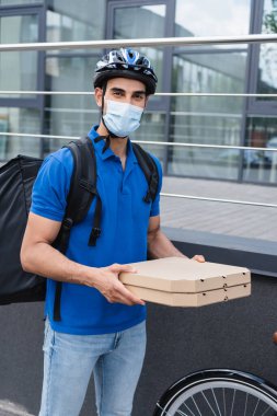 Tıbbi maskeli Müslüman kurye dışarıda bisiklet yakınında pizza kutuları tutuyor. 