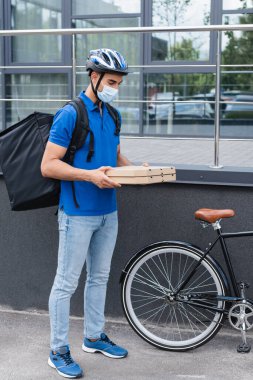 Koruyucu maskeli Arap kurye dışarıda bisiklet yanında pizza kutuları tutuyor. 