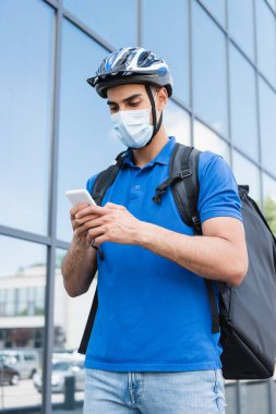 Kasklı ve tıbbi maskeli Müslüman kurye şehir caddesinde akıllı telefon kullanıyor. 