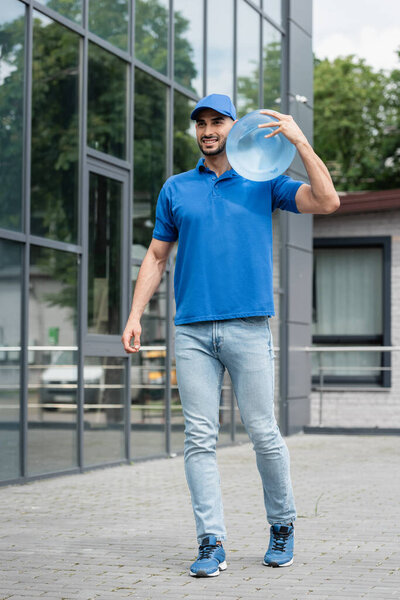 Cheerful muslim courier in uniform holding bottle of water outdoors 