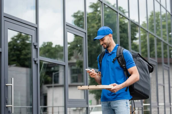 Lächelnder Muslimischer Kurier Mit Pizzakiste Und Smartphone Steht Neben Gebäude — Stockfoto