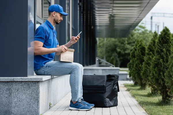 Πλευρική Άποψη Του Muslim Deliveryman Χρησιμοποιώντας Smartphone Και Κρατώντας Χάρτινη — Φωτογραφία Αρχείου