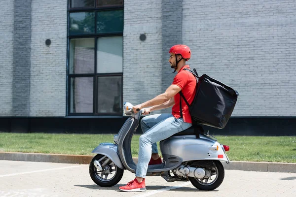 İslami kuryenin kırmızı üniformalı ve dışarıda scooter süren termo sırt çantalı görüntüsü. 