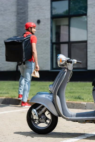 Scooter, termo sırt çantası ve kese kağıdıyla bulanık kuryenin yanında. 
