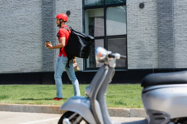 Smiling Arabian Deliveryman Coffee Paper Bag Walking Grass Blurred Scooter — Stock Photo, Image