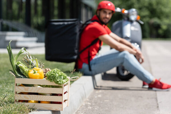 Коробка со свежими овощами рядом с размытым курьером и скутером на открытом воздухе 